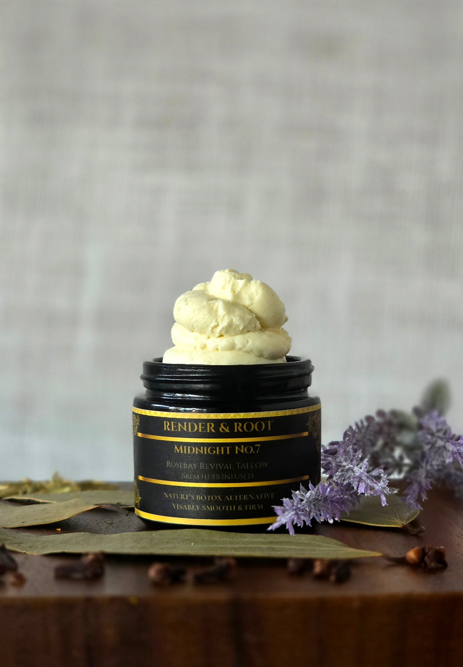 Whipped grass-fed beef tallow balm, Midnight No. 7, overflowing from a sleek 2oz black glass jar. Shown on a rustic wood surface surrounded by whole herbs, lavender, and dried botanicals to highlight our slow-infusion process. Rich, creamy texture for deep skin revival